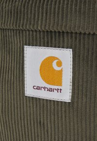 Carhartt-logo-merkki keltaisella emblemillä ja ruskealla tekstillä vihreässä ribbikorduroy-kankaassa.
