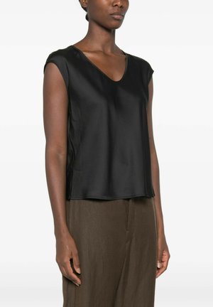 Vrouw draagt een mouwloze zwarte top met V-hals en losse bruine broek, staand tegen een effen witte achtergrond.
