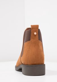 Bota Chelsea de ante marrón con paneles laterales oscuros de elasticidad y suela de goma negra, vista desde atrás sobre una superficie blanca.