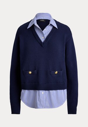 Πλεκτό πουλόβερ σε ναυτικό μπλε με λαιμόκοψη V, που φοριέται πάνω από λευκό και μπλε ριγέ πουκάμισο με χρυσά κουμπιά στις τσέπες του πουλόβερ.