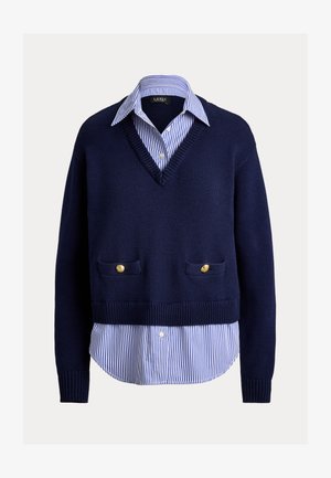 Πλεκτό πουλόβερ σε ναυτικό μπλε με λαιμόκοψη V, που φοριέται πάνω από λευκό και μπλε ριγέ πουκάμισο με χρυσά κουμπιά στις τσέπες του πουλόβερ.