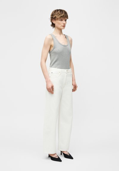 Νεαρό άτομο με κοντά μαλλιά, φορώντας γκρι αμάνικο τοπ, λευκό παντελόνι φαρδύ στο κάτω μέρος και μαύρα μυτερά τακούνια, στέκεται μπροστά σε απλό φόντο.