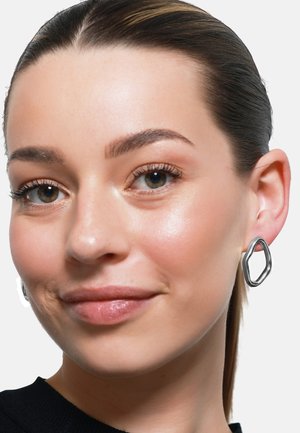 Silberne, strukturierte, geometrische Ohrringe in einer gewellten, achteckigen Form; das Modell zeigt glänzende Haut und dunkles Haar in einem schwarzen Hemd.