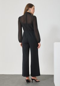 Black jumpsuit with sheer long sleeves, high neck, and fitted waist. Wide-legged pants, worn with black heels. Minimalist style.