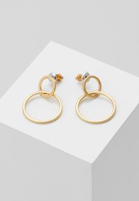 Zlaté náušnice se dvěma propletenými otevřenými kruhy, menším nahoře a větším pod ním, vystavené na bílém podstavci.