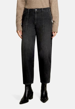 Person wearing high-waist black cropped jeans with large front pockets, beige sweater, and black heeled ankle boots on a neutral background.