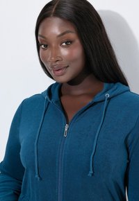 Felpa blu con zip e cordoncino, caratterizzata da una morbida texture e un design aderente. Le cuciture con zip aggiungono funzionalità al capo.