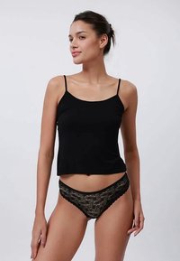 Young woman with dark hair tied back wearing a black tank top and black lace underwear, standing against a plain light background.