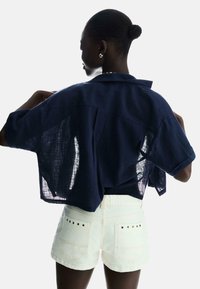 Tengerészkék vászon ing, laza, cropped fazonnal és rövid ujjakkal, fehér farmer sorttal párosítva, amelyen szegecselt részletek találhatók a hátsó zsebeknél.