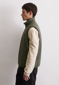 Zielona, bezrękawnikowa kamizelka z gładkiego materiału, z wysokim kołnierzem, zestawiona z beżową, teksturowaną bluzką na długi rękaw oraz czarnymi spodniami.