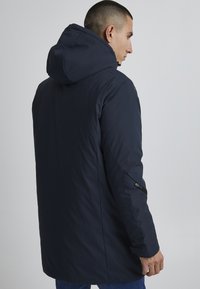 Marineblå parka-jakke med hette, som har en glidelåslomme på siden og en jevn, matt tekstur. Designet for varme og komfort.