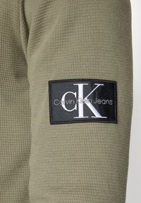 Tissu côtelé vert olive avec un patch rectangulaire noir portant le texte blanc "CK" et "Calvin Klein Jeans." Détails de surface texturée visibles.