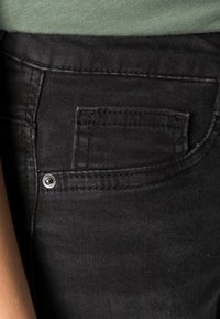 Jeans de denim negro con un detalle en el bolsillo trasero, que cuenta con un remache plateado. La tela tiene una textura suave.