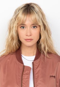 Chaqueta bomber color rosa con cremallera frontal, cuello acanalado y logotipo bordado. Llevada sobre una camiseta blanca, que presenta una textura suave y brillante.