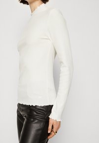 Top bianco a maniche lunghe con collo alto e orlo ondulato, abbinato a pantaloni in pelle nera. La texture è liscia e aderente.