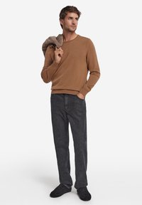 Brun tröja i mjuk textur, kombinerad med mörkgrå jeans. Modellen håller ett ljusbrunt plagg över axeln och bär svarta skor.