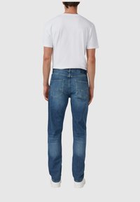 Jean en denim bleu pour homme avec une coupe droite, présentant un lavage moyen, des poches arrière et une ceinture standard. Assorti avec un t-shirt blanc.