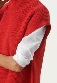 Rode gebreide mouwloze top met een hoge hals, voorzien van horizontale ribbels. Gelaagd over een witte langarmshirt, met zichtbare manchetten.