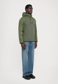 Sweatshirt com capuz em verde oliva com um padrão texturizado e logótipo preto, emparelhado com jeans de perna reta em azul claro e sapatos pretos.