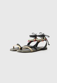 Sandalias de cuero negro con acentos dorados, cintas en el tobillo y cuentas decorativas que presentan elementos en verde, rojo y blanco. Diseño de suela plana.