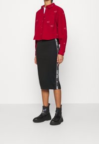 Červená zkrácená košile na knoflíky s vyšívaným logem, kombinovaná s černou pouzdrovou sukní s bílými pruhy po stranách a černými platformními kotníkovými botami.