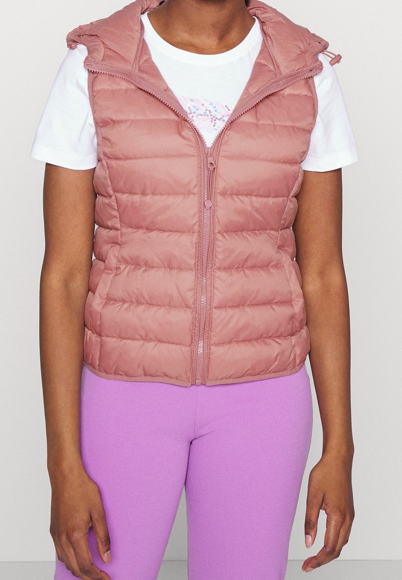 Personne portant un gilet matelassé sans manches rose sur un T-shirt blanc et des leggings violets, debout avec les bras détendus le long du corps.