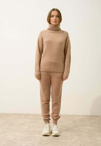 Beige neulepaita, jossa on korkea kaulus, yhdistettynä samanvärisiin kapeneviin housuihin, molemmat pehmeää materiaalia. Asusteena vaaleat urheilukengät.