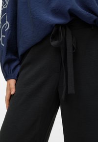 Μπλε ναυτικό κορυφή με υφή και φαρδιές μανίκια, συνδυασμένη με μαύρο παντελόνι στη μέση με δέσιμο και απαλό ύφασμα.