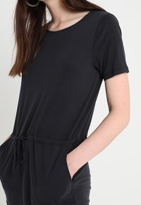 Combinaison noire à manches courtes, décolleté arrondi, taille élastique et poches latérales. Tissu doux au toucher lisse.