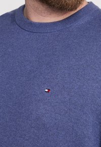 Maglione blu lavorato a maglia con scollo rotondo e polsini a coste. Presenta un piccolo logo rosso, bianco e blu sul petto. Texture morbida.