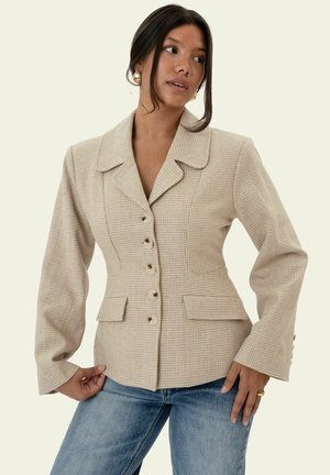 Frau trägt beigen, maßgeschneiderten, geknöpften Blazer mit weitem Kragen und blaue Jeans, posiert mit den Händen in der Hüfte und schaut zur Seite.