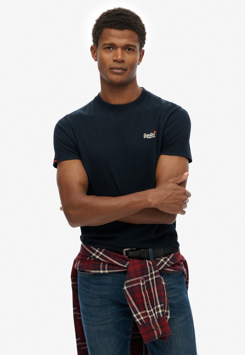 Camiseta de algodón azul marino con cuello redondo y mangas cortas; presenta un pequeño logo en el pecho. Lleva una camisa de franela a cuadros anudada a la cintura.