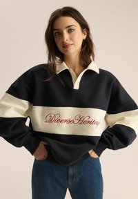 Granatowy i kremowy sweatshirt z kołnierzykiem polo, z haftem "Diverse Heritage" na czerwono na klatce piersiowej. Miękki, gruby materiał.