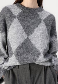 Nainen, joka käyttää harmaata ja vaaleanharmaata vinoruutukuosisia villapaitaa, joka on laitettu korkeavyötäröisten tummanharmaiden pleatattujen housujen sisään.