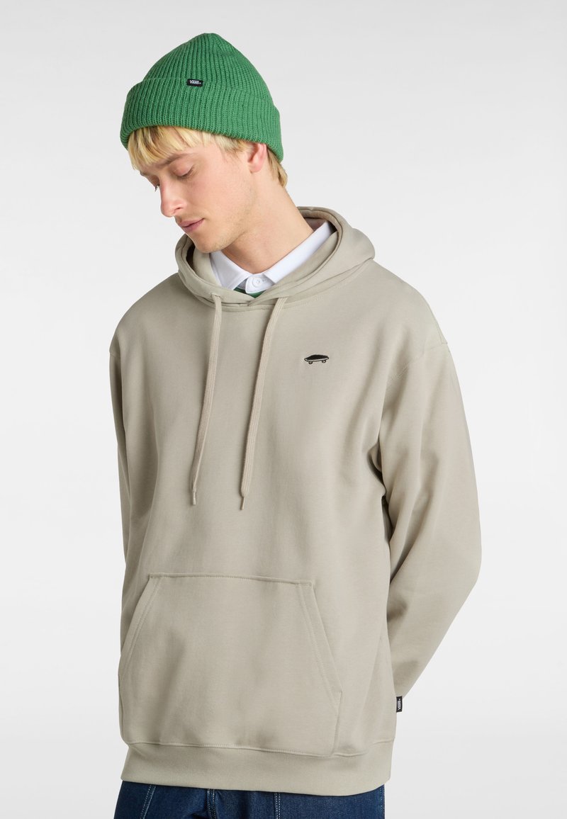 Ljusbeige hoodie med framficka, dragskor och liten svart skateboardlogga. Tillsammans med en grön ribbad stickad mössa.