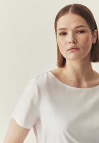 Haut blanc à manches courtes avec un col rond, une texture lisse et un design minimaliste, présenté sur un fond clair.