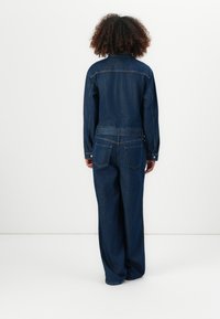 Donkerblauwe denim jumpsuit met lange mouwen, een kraag en een wijduitlopende pasvorm. Opvallende achterzakken en gestikte details.
