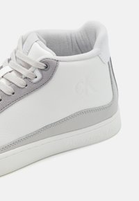 Balta ādas sneakers ar pelēku zamšādas akcentiem, apaļš pirksts, teksturētas virsmas, kontrastējošas šuves un iegriezts logotips. Izlīdzinātas šņorītes un mīksts apkaklīte.