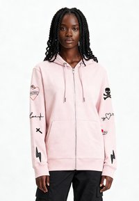 Felpa con cappuccio rosa con zip, realizzata in cotone, con toppe nere ricamate sulle maniche che includono cuori, teschi e fulmini.