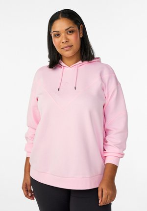 Vrouw die een lichtroze hoodie met trekkoorden draagt, zwarte broek, staand tegen een effen witte achtergrond, naar voren kijkend met een neutrale uitdrukking.