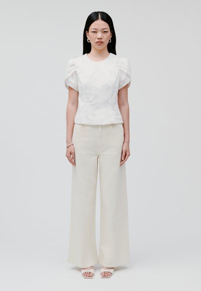 Top bianco con maniche a sbuffo in tessuto testurizzato, abbinato a pantaloni beige a vita alta e a gamba larga e sandali beige con cinturini.