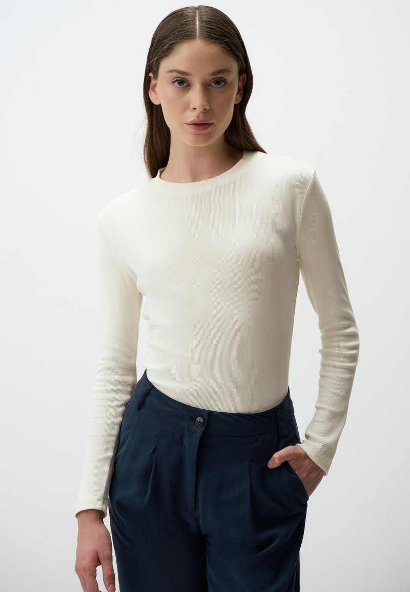 Maglietta a maniche lunghe in crema con costine abbinata a pantaloni navy a vita alta, caratterizzati da un dettaglio con bottone e tasche laterali. Design pulito e testo liscio.
