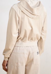 Béžová mikina s kapucí z měkkého materiálu a žebrovaným lemem, v kombinaci s volnými krémovými kalhotami se zadními kapsami.