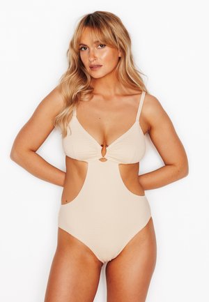 Donna con lunghi capelli biondi che indossa un costume intero beige con ritagli, in piedi con le mani dietro la schiena contro uno sfondo bianco.