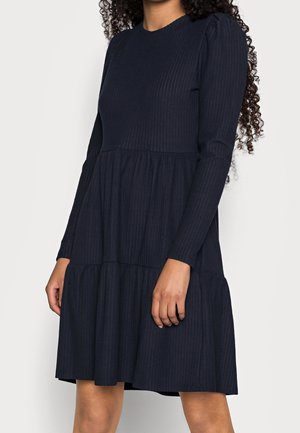 Kvinde iført en langærmet, ribstrikket mørkeblå kjole med en lagdelt nederdel, stående mod en ensfarvet hvid baggrund.