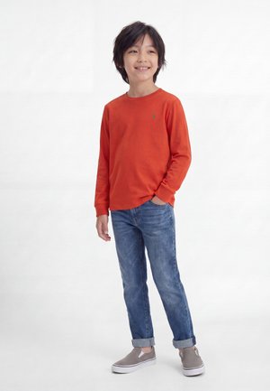 Unge dreng iført en orange langærmet skjorte, blå jeans med manchetter og grå slip-on sko, står med den ene hånd i lommen og smiler.