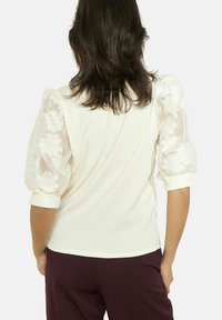 Puhvis varrukatega topp, pehme kreemika kangaga ja õrna, lillemustriga tikandiga läbipaistvate varrukatega; kehale vormiv lõige, millel sooniline tekstuur.