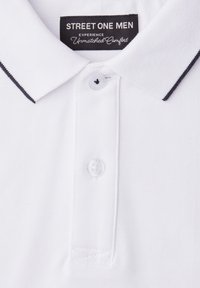 Polo pour homme blanc avec bordure noire sur le col, deux boutons blancs, et une étiquette noire portant l'inscription "STREET ONE MEN Experience Unmatched Comfort."