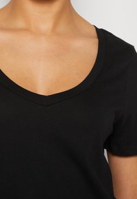Nærbillede af en person iført en sort V-hals T-shirt med korte ærmer, der viser nakke, kraveben og øverste brystområde på en hvid baggrund.