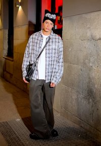 Prevelik sivi kariran srajc na belem T-majici, temno široke hlače, črna kapa z belim vzorcem in črna torba čez ramo.